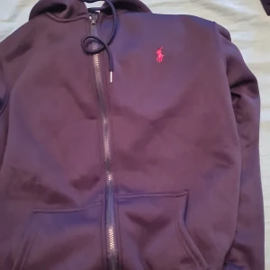 Mörkblå hoodie från Polo Ralph Lauren - Säljer en snygg mörkblå hoodie från Polo Ralph Lauren med dragkedja och broderad röd logga på bröstet. Perfekt för en stilren och avslappnad look. 🧢