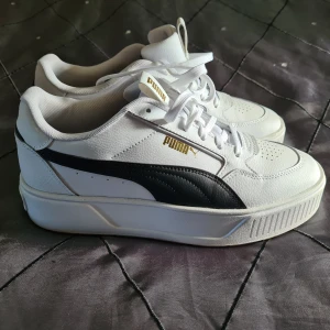 Vita sneakers från Puma - Snygga vita sneakers från Puma med svarta detaljer och guldlogga. Skorna har en klassisk design med snörning och en bekväm sula. Perfekta för en stilren look.