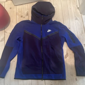 Blå och svart jacka från Nike - Säljer en blå och svart jacka från Nike med dragkedja och huva. Jackan har en sportig design med Nike-logga på bröstet och praktiska fickor. Perfekt för en aktiv livsstil.