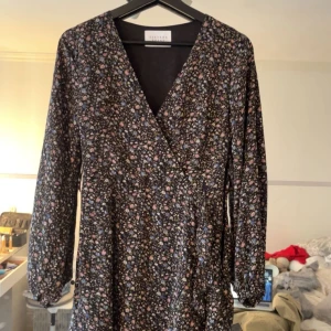 Blommig klänning från Sisters Point - Säljer en söt blommig klänning från Sisters Point. Klänningen har en v-ringad design och långa ärmar, perfekt för både vår och höst. Den har ett fint blommönster i olika färger på en mörk bakgrund.