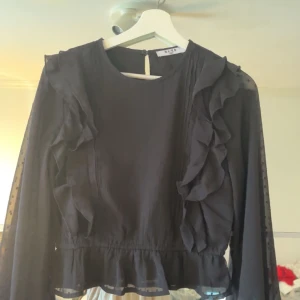 Svart blus med volangdetaljer - Elegant svart blus från Next med volangdetaljer framtill och genomskinliga prickiga ärmar. Perfekt för en stilfull look.