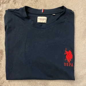 Mörkblå t-shirt från U.S. Polo Assn. - Säljer en mörkblå t-shirt från U.S. Polo Assn. med röd broderad logga på bröstet och en stor siffra på ärmen. T-shirten har en klassisk rund hals och korta ärmar. Perfekt för en avslappnad stil.
