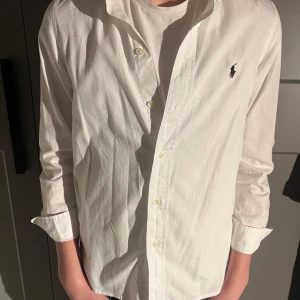 Vit och blå skjorta från Ralph Lauren - Säljer två klassiska skjortor från Ralph Lauren. En vit och en blå, båda med den ikoniska loggan broderad på bröstet. Skjortorna har långa ärmar och knappar framtill. Perfekta för en stilren look!
