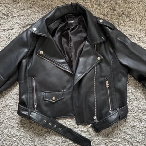 Svart skinnjacka från Pull&Bear - Snygg svart skinnjacka från Pull&Bear med dragkedjor och bälte. Jackan har en klassisk bikerstil med långa ärmar och flera fickor med dragkedja. Kort modell, tyvärr slitning vid öglan för att hänga upp jackan, annars jättefint skick då den endast är använd 2-3 gånger☺️