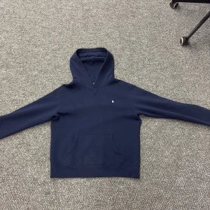 Mörkblå hoodie från Ralph Lauren - Säljer en stilren mörkblå hoodie från Ralph Lauren med det klassiska logotypbroderiet på bröstet. Tröjan har en känguruficka och en bekväm huva. Perfekt för en avslappnad look.