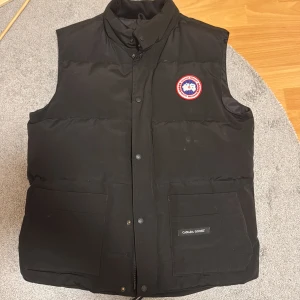 Svart dunväst från Canada Goose - Säljer en svart dunväst från Canada Goose med klassisk logga på bröstet. Västen har en hög krage och stängs med både dragkedja och knappar. Två stora fickor framtill ger extra funktionalitet. Perfekt för kyliga dagar. Storleken är XL fast skulle ändå passa en person som har Storlek L på jackor/västar. Mycket fräsch och endast använd 2-3 gånger.