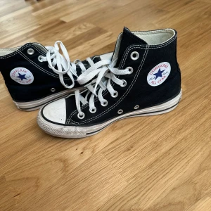 Converse - Klassiska svarta Converse All Star sneakers i hög modell med vit sula och snörning. Skorna har det ikoniska Converse-märket på sidan och är tillverkade i canvasmaterial. Perfekta för en avslappnad stil.