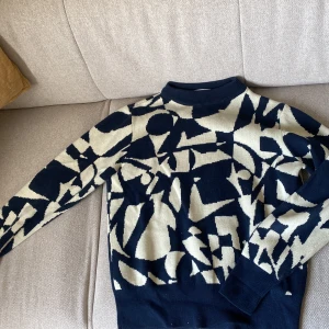 Mönstrad stickad tröja från Monki - Säljer en cool stickad tröja från Monki i storlek XS. Tröjan har ett abstrakt mönster i mörkblått och beige, med ribbade muddar och hög hals. 