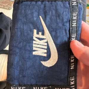Blå Nike plånbok - Snygg blå plånbok från Nike med klassisk broderad logga och svart kant med Nike-tryck. Plånboken har flera fack för kort, myntficka med dragkedja och ett transparent fack. Perfekt för att hålla koll på dina grejer.