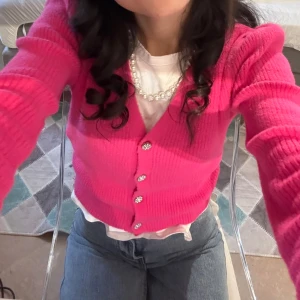 Rosa kofta från Zara med pärlknappar - Superfin rosa kofta från Zara med v-ringning och glittriga pärlknappar. Koftan är ribbstickad, har lång ärm och är croppad i modellen. Perfekt att styla med jeans eller kjol för en trendig look.