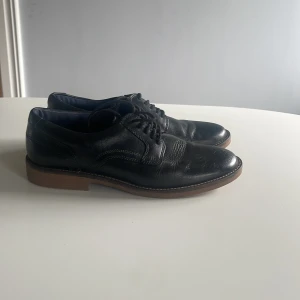 Svarta skinnloafers med snörning - Klassiska svarta loafers i skinn med snörning och brun sula. Snyggt stilrena med rund tå och diskreta sömmar, perfekta till både jeans och kostym. Skorna har en platt sula och tidlös design som funkar året runt. Kan hämtas upp i Huddinge annars står man för frakten själv!