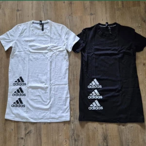 Adidas t-shirtklänningar svart & vit - Två t-shirtklänningar från Adidas, en i vitt och en i svart. Båda har rund halsringning, korta ärmar och tre stora Adidas-loggor tryckta längs ena sidan. Skön bomullskänsla och avslappnad passform, perfekt för en sportig look.