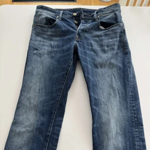 G-Star Raw 3301 Straight jeans blå - Säljer ett par klassiska blå jeans från G-Star Raw, modell 3301 Straight. Jeansen har raka ben, fem fickor och snygga slitningar framtill. De är i denim med kontrastsömmar och har en normal passform. Perfekta till vardagslooken!