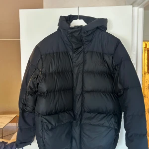 Svart dunjacka från The North Face - Säljer en svart dunjacka från The North Face med huva och stora fickor framtill. Jackan är vadderad och har en klassisk puffer-look med horisontella sömmar. Perfekt för kalla dagar och riktigt snygg till streetwear-stilen.