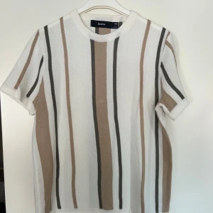 Randig stickad t-shirt från Bershka - Säljer en stickad t-shirt från Bershka med vertikala ränder i vitt, beige och grått. Tröjan har korta ärmar, rund halsringning och en relaxed fit. Perfekt för dig som gillar en chill och stilren look.