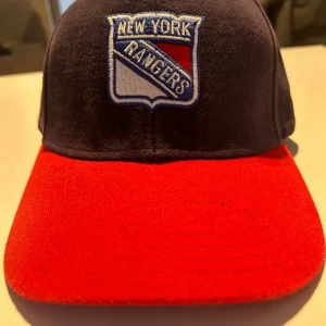New York Rangers keps  - Snygg keps med New York Rangers-logga framtill och NYR broderat bak. Kepsen är mörkblå med röd skärm och justerbar rem baktill. Perfekt för hockeyfans som vill visa sitt lagstöd.
