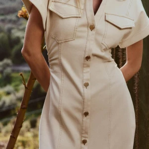 Beige skjortklänning med knappar - Snygg beige jeansklänning med korta ärmar och guldiga knappar hela vägen framtill. Klänningen har två bröstfickor med lock och markerad midja för en smickrande siluett. Perfekt för en trendig och avslappnad look.Använd 1 gång 