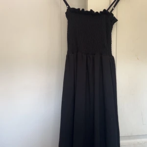 Svart smockad långklänning med volang - Svart långklänning med smockad överdel, tunna justerbara axelband och söt volangkant upptill. Kjolen är luftig och faller snyggt. Perfekt för dig som gillar en enkel men stilren look.