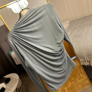 Ljusblå långärmad topp med drapering - Säljer en ljusblå långärmad topp med snygg drapering och rynkade detaljer över ryggen. Tunt och mjukt material som känns skönt mot huden. Perfekt för dig som vill ha något unikt och stilrent i garderoben.