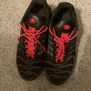 Nike Air Max Plus Tn Svart/Röd - Nike Air Max Plus Tn sneakers i svart med röda detaljer och snörning. Skorna har vågformade mönster på ovandelen, röd swoosh på sidan och Tn Air-logga bak. Snygg och sportig design med robust sula och bekväm passform. Perfekta för dig som gillar streetwear.