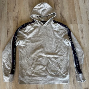 Grå hoodie från GANT med logoband - Snygg grå hoodie från GANT med huva och känguruficka. Svarta logoband med GANT-tryck längs ärmarna ger en sportig vibe. Mjuk bomullskänsla och relaxed fit, perfekt för chill dagar. Klassisk look med moderna detaljer.