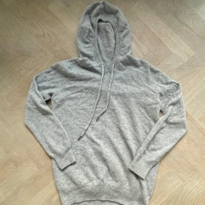 Ljusgrå hoodie med snörning - Mysig ljusgrå bland kashmir hoodie med huva och snörning. Tröjan är långärmad och har ribbade muddar vid ärmslut och nederkant. Tillverkad i mjukt stickat material som känns skönt mot huden. Perfekt för chill dagar eller när du vill ha en enkel och stilren look. Tveka inte på att höra av er!!!