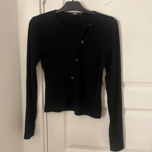 Svart ribbad kofta från BikBok - Svart ribbad kofta från BikBok i storlek M. Snygg asymmetrisk knäppning framtill med flera knappar och långärmad modell. Säljs för 50kr
