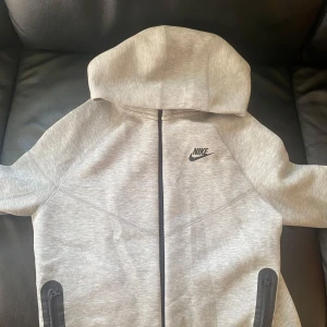 Grå Nike hoodiejacka XL (158-170cm) - Snygg grå hoodiejacka från Nike i storlek XL(158-170cm. Jackan har dragkedja framtill, huva och svarta detaljer vid fickorna. Perfekt för en sportig look och chill vardagsstil. Tillverkad i mjukt material som känns skönt mot huden. Nike-logga på bröstet.