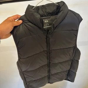 Svart dunväst från Uniqlo - Säljer en svart, quiltad dunväst från Uniqlo i modellen Ultra Light Down. Västen har hög krage och dragkedja framtill. Den är lätt och smidig, perfekt att slänga över en hoodie eller tröja när det är kyligt ute.