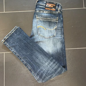 Jack & Jones Slim Glenn jeans blå - Snygga blå jeans från Jack & Jones, modell Slim Glenn. Just denna modellen går inte att få tag på längre vad jag kan se så den är exclusiv. Jeansen har en slim passform, klassisk femficksdesign och coola slitningar framtill. De är tillverkade i jeansmaterial med orangea detaljer och kontrastsömmar. Perfekta för en trendig och avslappnad look. 