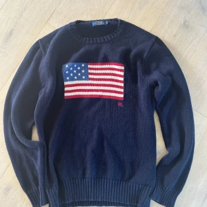 Mörkblå stickad tröja Polo Ralph Lauren - Mörkblå stickad tröja från Polo Ralph Lauren med amerikansk flagga broderad på bröstet och RL-detalj. Klassisk rund halsringning och ribbade muddar. Perfekt för dig som gillar preppy stil och vill sticka ut med en ikonisk look.