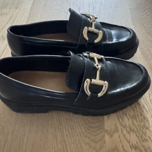 Svarta loafers med gulddetaljer - Klassiska svarta loafers i skinn med snygga guldfärgade metalldetaljer framtill. Skorna har rund tå och platt sula, vilket gör dem både stilrena och bekväma. Perfekta för dig som gillar en tidlös och trendig look.