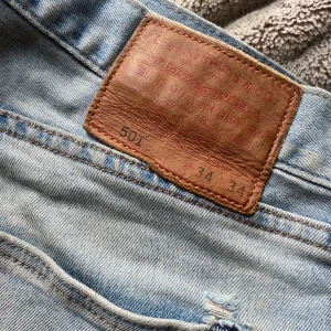 Levi's 501 ljusblå jeans W34 L34 - Säljer ett par klassiska ljusblå Levis 501 jeans med raka ben och låg midja. Jeansen har en avslappnad passform och är tillverkade i slitstarkt denim. Köpt på herravdelningen men vem som kan bära dem.  Hål på knät och är W34 och L34. Perfekta för en cool och avslappnad stil, förförallt för den längre personen. Finns en liten fläck vid fickan. 