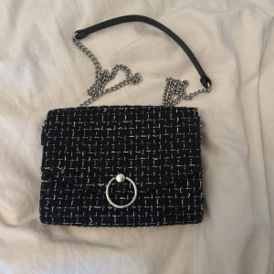 Svart axelväska med kedja och ringdetalj - Stilren svart axelväska i rutigt tyg med silverfärgad kedjerem och en snygg ringdetalj på locket. Väskan har ett mjukt innerfoder och stängs med magnetknapp. Perfekt storlek för mobil och småsaker, och ger en cool touch till din outfit.