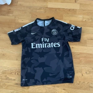 PSG Nike svart camo fotbollströja S - Snygg svart Paris Saint-Germain fotbollströja från Nike med vit logga och Fly Emirates-tryck. Tröjan har korta ärmar, camo-mönster och är tillverkad i Dri-FIT-material som andas. Perfekt för dig som vill sticka ut på planen eller på läktaren.