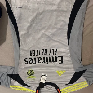 Real Madrid målvaktströja Adidas - Snygg benfica tröja från Adidas i ljusgrått med svarta detaljer och neongula inslag. Tröjan har lång ärm, Aeroready-material och klubbmärke samt sponsortryck på bröstet. Perfekt för dig som vill sticka ut på planen.