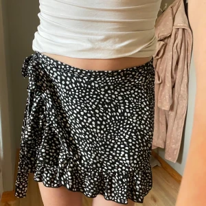 Svart omlottkjol med vita prickar - Supersöt svart kort omlottkjol med vita prickar och volangkant. Kjolen knyts i sidan med en rosett och har en flowy känsla som är perfekt för sommardagar. Lätt och luftig, passar perfekt till en enkel topp eller hoodie. Shorts intill som är perfekt då de blåser💕