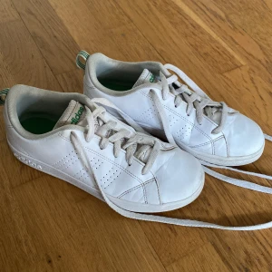 Vita sneakers från Adidas med gröna detaljer - Klassiska vita sneakers från Adidas med gröna detaljer på plösen och hälen. Skorna har snörning, perforerade sidor och rund tå. Ovandelen är i skinn och insidan är fodrad i textil. Perfekta för en clean och sportig look.