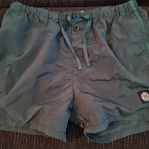 Gröna badshorts från Stone Island.Helt nya! - Snygga gröna badshorts från Stone Island med klassisk logga på benet. Shortsen har elastisk midja med snörning och knappar framtill, samt en bakficka. Perfekta för stranden eller poolen, riktigt clean look och skön passform.