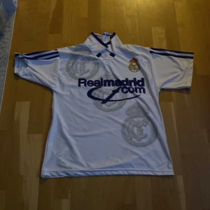 Real Madrid Figo 10 fotbollströja - Klassisk vit Real Madrid-tröja med blå detaljer, Figo 10 på ryggen och klubbmärke på bröstet. Adidas-logga och RealMadrid.com som sponsor framtill. Snygg krage med blå ränder på ärmarna. Perfekt för dig som älskar retro och fotbollshistoria.