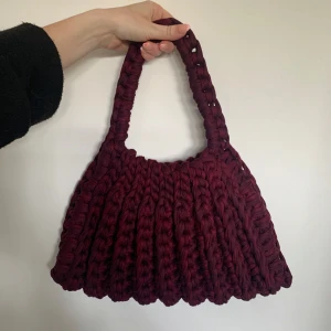 Vinröd virkad axelväska - Superfin vinröd virkad axelväska. Väskan har en mjuk, ribbad struktur och breda handtag som gör den både snygg och bekväm att bära. Perfekt accessoar för att lyfta din outfit med en unik touch.