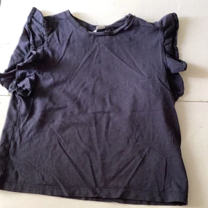 Mörkblå topp med volangärmar från Gina Tricot - Säljer en mörkblå topp från Gina Tricot med volangdetaljer på ärmarna. Toppen har rund halsringning och är tillverkad i mjukt material, perfekt för en stilren och enkel look.