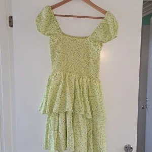 Gul blommig kortklänning med volanger - Supersöt gul kortklänning med små vita blommor, puffärmar och smockad överdel. Klänningen har volanger i kjolen och är perfekt för sommarens alla tillfällen. Aldrig använd. Lite mer gul i verkligheten än den är på bilden!💕