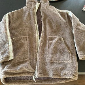 Beige teddyjacka i storlek XL - Mysig och oversized teddyjacka i beige med stora fickor framtill och dragkedja. Jackan har vita detaljer längs ärmarna och är fodrad med quiltat innerfoder för extra värme. Perfekt för kyliga dagar när du vill vara både bekväm och snygg.
