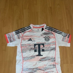 Bayern München Adidas fotbollströja - Snygg Bayern München fotbollströja från Adidas med korta ärmar. Tröjan är vit med grå och rosa mönsterdetaljer, svarta Adidas-ränder på axlarna och klubbmärke på bröstet. Stort T-Mobile-tryck på framsidan och patch på ärmen.