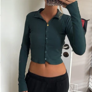 Mörkgrön croppad skjorta med knappar - Snygg mörkgrön croppad skjorta med långa ärmar och knappar framtill. Skjortan har en klassisk krage och sitter tight mot kroppen, perfekt för en trendig look.