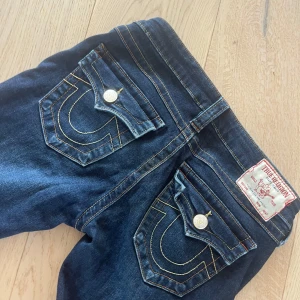 True Religion jeans låg midja blå - Säljer ett par snygga True Religion jeans i mörkblå denim med kontrastsömmar och ikoniska bakfickor med lock och stora knappar. Jeansen har låg midja och klassisk femficksdesign. Perfekta för dig som gillar street och premium denim. Dem har knappar på insidan för att kunna spänna/lösa upp midjan. Det enda som är att tänka på  är att  själva loggan på baksidan som har lite lösa sömmar. Priset kan diskuteras!! Hoppas ni finner dem intressanta!!💕