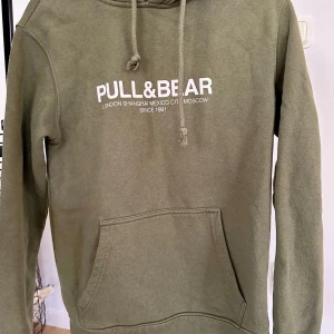 Olivgrön hoodie från Pull&Bear XS - Olivgrön hoodie från Pull&Bear med vit logga framtill. Klassisk känguruficka, snörning i huvan och mjukt material. Perfekt för chill dagar eller när du vill ha en enkel och snygg look.