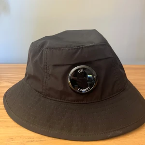 Svart bucket hat från C.P. Company - Stilren svart bucket hat från C.P. Company med den ikoniska linsen på sidan. Hatten är tillverkad i lätt och slitstark polyester, perfekt för att lyfta din streetwear-look. Klassisk form med bred brätte och diskret sömnadsdetalj runt om. Använd två gånger, som ny!