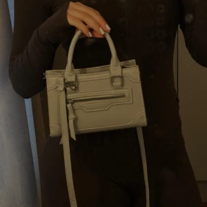 Grå handväska med dragkedja - Snygg grå handväska i skinnimitation med både handtag och axelrem. Väskan har en stilren design med dekorativa sömmar och en praktisk dragkedja framtill. Perfekt storlek för det viktigaste och passar till många outfits.